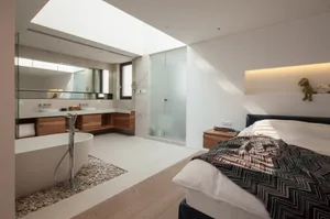 Modern bedroom featuring white walls, a bed with modern bedding, a floating vanity, and a stone-lined bathtub.