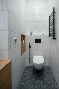 A modern bathroom with white tiles and black terrazzo flooring, featuring a wall-mounted toilet with a black cistern, wooden 