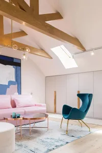 A stylish, modern living room with a pink sofa, blue chair, wooden beams, and a skylight.