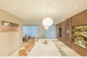 Modern living and dining room with wooden chairs, glass windows, and a white pendant light hanging above a long dining table.
