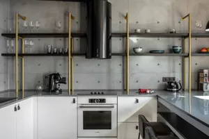 A modern minimalist kitchen with concrete walls, featuring a black and gold shelving unit, white cabinetry, and a sleek oven.