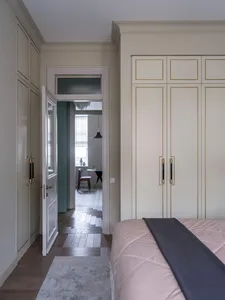 A minimalist bedroom featuring a bed with a pink blanket, a white wardrobe with gold trim, and a wooden floor with a gray rug