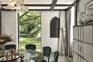 A modern minimalist dining room featuring a glass table surrounded by green velvet chairs, with a view of a lush garden throu