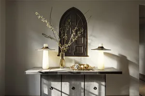 A ceramic decorative table with a vase of flowers, two lamps, and decorative objects on top of it, against a wall with a wind