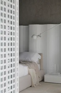 A minimalist bedroom featuring a white bed with a beige throw, a white lamp mounted on the wall, and a white perforated scree