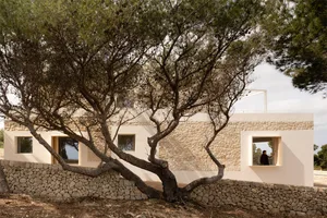 Photo of a modern residential house with stone walls, featuring large windows and a person standing near one of the windows.