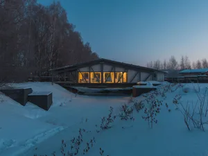 A modern winter cabin with warm lights illuminated through large windows amidst snowy surroundings.