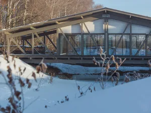 A modern floating house with a metal frame and transparent glass walls, surrounded by snow-covered ground and leafless trees 