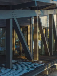 Modern wooden porch with sunlight shining through beams and windows, featuring a wooden deck covered in snow.