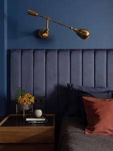 A modern bedroom featuring a dark blue velvet headboard, brass wall lamp, wooden nightstand, and orange throw pillows.