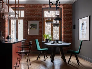 A modern kitchen featuring exposed brick walls, a round dining table with green chairs, and a bar counter with hanging glassw
