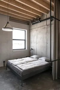 A minimalist bedroom with a concrete floor, white bedding, and industrial-style wood beams on the ceiling.