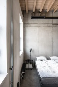A minimalist bedroom with concrete walls and floor, featuring a white bed with white pillows, a black side table with a black