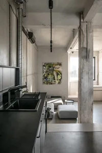 Modern minimalist living room with concrete and steel elements, featuring a kitchen area with black countertops and industria