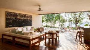 Modern living room with white sofas, cushions, and wooden furniture surrounded by large windows overlooking a lush green land