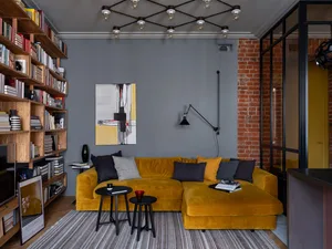 A modern living room featuring a yellow velvet sofa, black stools, and a large bookshelf.