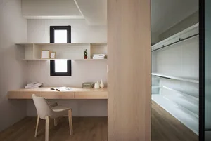 A modern minimalist home office featuring a wooden desk and shelves, with a comfortable chair and a small window.