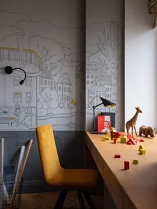 A modern playroom featuring a wooden desk with toys and a mustard yellow chair, against a wall mural depicting a small villag