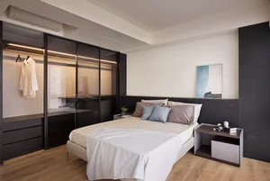 A modern bedroom with minimalist lighting, featuring a bed with white bedding and multiple pillows, a glass wardrobe, and woo