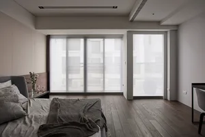 A modern bedroom with white walls, wooden floor, and a large window with blinds, featuring a bed with a gray blanket and cush