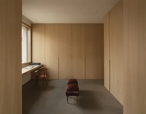 An open, minimalist living area with wooden cabinetry, a bench, and a small desk area by a window.
