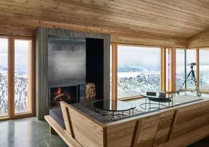A modern living room with a wooden sectional sofa, glass tables, and a fireplace with a view of snowy mountains.