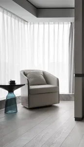 A modern residential space featuring a beige upholstered chair with a glass table and sheer curtains by the window.