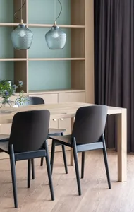 A modern dining room featuring black chairs around a wooden table, with a wooden floor and a shelving unit in the background.