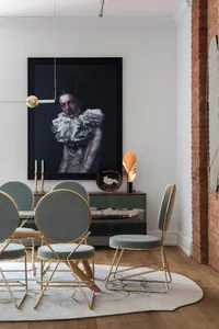 A modern dining room features metallic chairs with blue velvet upholstery, a glass table, and a large portrait of a woman.