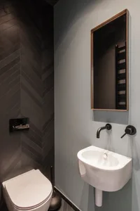 Modern minimalist bathroom with black chevron-patterned tiles, white appliances, and a wall-mounted sink with brass faucet an