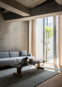 A modern, minimalist living room featuring a concrete wall, glass coffee table, and large floor-to-ceiling windows.