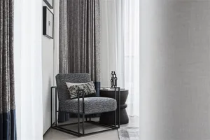 A modern minimalist living room featuring a grey upholstered chair with metal frame, white curtains, and a black side table.