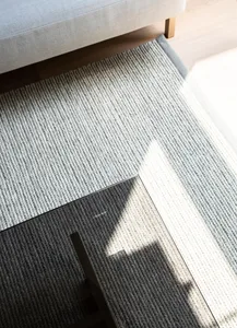 A close-up view of a modern grey knit rug with a sunlit pattern, placed under a grey cushioned sofa on a light wooden floor.