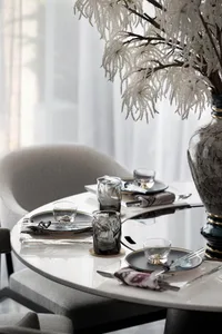 Modern dining room featuring sleek glass tables and chairs, with white curtains and decorative white flowers in a vase.
