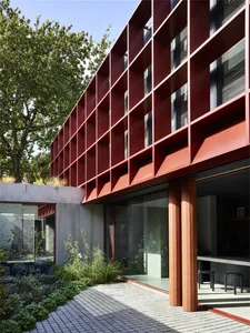 Modern metal-frame residential building exterior with red accents and a green courtyard surrounded by greenery.