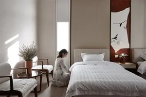 A minimalist bedroom with a white fabric bed, a plant, and a lamp.