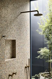 Industrial bathroom featuring a terrazzo shower wall, brass fixtures, and a large window with green plants outside.