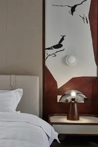 A minimalist bedroom featuring white furniture and a white headboard with a wooden base, complemented by a white table lamp o