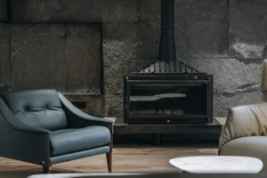 A modern living room featuring a concrete fireplace, teal armchair, and floor-to-ceiling stone wall