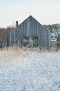 A minimalist wooden house surrounded by snow-covered grass and trees in a winter landscape.