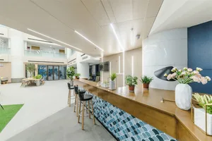 Modern lobby area with a bar counter, chairs, and greenery. Light blue and white interior design.