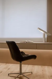 A modern workstation featuring a black leather chair, a table with a table lamp, and a window with blinds.