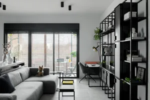 A modern minimalist living room featuring a grey sectional sofa, a glass coffee table, and a black and white shelving unit wi