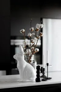 A modern white ceramic bunny sitting on a marble surface with a glass vase containing cotton branches and black candle holder