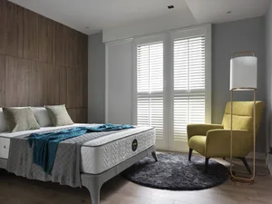 A modern bedroom with wooden accents, featuring a grey bed and yellow armchair, illuminated by natural light through white sh
