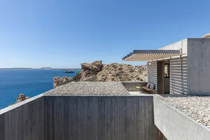 A modern, concrete house built into a rocky cliffside overlooking the ocean.
