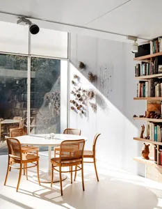 A modern dining room with a white table, wooden chairs, and glass wall providing a view of the outdoors.