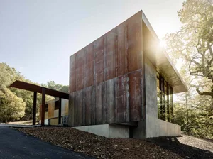 A modern metal-clad house with large windows and concrete base in a wooded area.