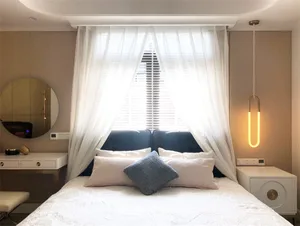 A modern bedroom featuring a bed with a white bedspread and blue pillow, white curtains, and a white headboard.