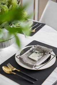 Modern dining table set with glass plates, metal cutlery, greenery centerpiece, and place mats.